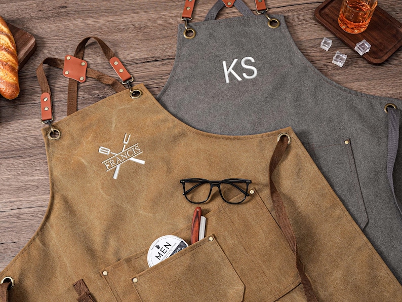 May include: Two aprons, one brown and one gray, with leather straps and pockets. The brown apron has the name "FRANCIS" and a crossed fork and spatula logo. The gray apron is personalized with the initials "KS". Black eyeglasses and a pen rest in the brown apron pocket.