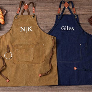 May include: Two aprons, one brown and one navy blue, with leather straps and pockets. The brown apron has the initials "NK" embroidered on it, while the blue apron has the name "Giles". Both aprons are designed for kitchen or workshop use.