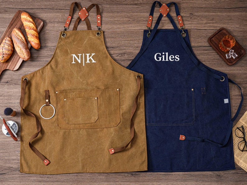 May include: Two aprons, one brown and one navy blue, with leather straps and pockets. The brown apron has the initials "NK" embroidered on it, while the blue apron has the name "Giles". Both aprons are designed for kitchen or workshop use.