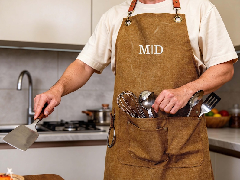 May include: Brown apron with leather straps and metal hardware. The apron has two pockets and the initials "MID" embroidered on the front. A spatula is held in one hand, while cooking utensils are in the pocket.