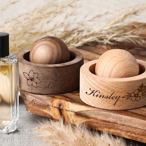 Può includere: Una bottiglia di profumo in vetro trasparente con un tappo nero accanto a due oggetti decorativi in legno. Uno è marrone scuro con un motivo floreale e la parola "dove" incisa. L'altro è in legno chiaro con il nome "Kinsley" inciso.