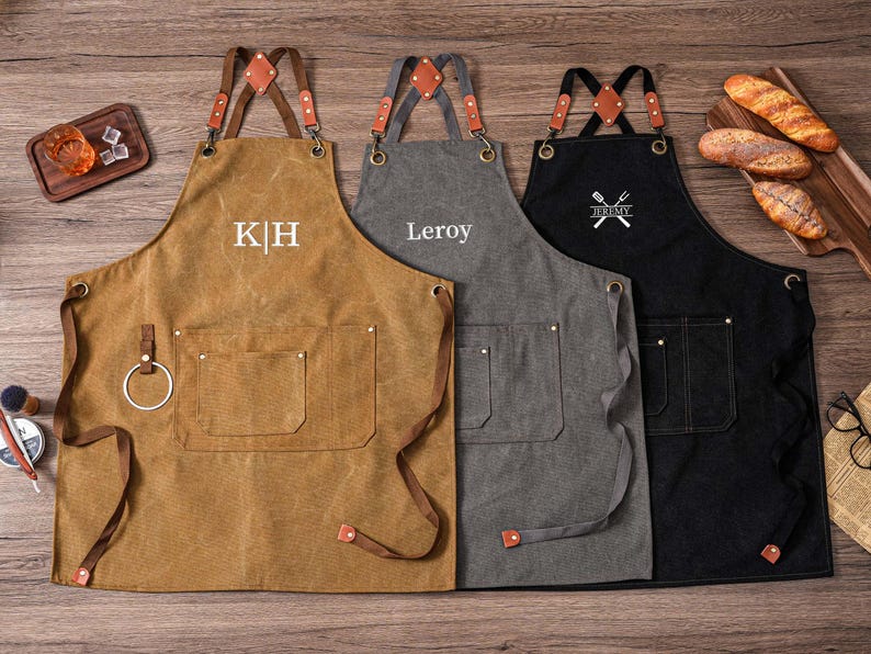 May include: Three personalized aprons in brown, gray, and black, with leather straps and pockets. The brown apron has the initials "K H", the gray apron has the name "Leroy", and the black apron has a crossed fork and knife design with the name "Jeremy".