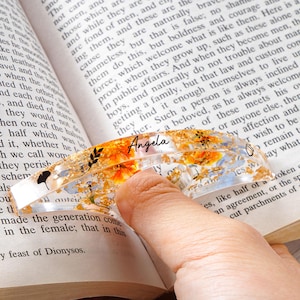 May include: Clear resin bookmark with gold flakes, orange flowers, and black leaf accents. The bookmark has the name "Angela" in black script. It is placed on an open book.