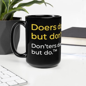 Op de afbeelding: Zwarte keramische mok met een witte en gele inscriptie die luidt "Doers do but don't Don'ters do but do. TM"