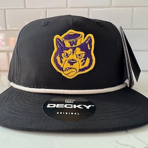 Puede incluir: Gorra de béisbol negra con visera plana y detalle de cuerda blanca. La gorra presenta un parche de un perro morado y dorado con un sombrero. La marca "DECKY ORIGINAL" es visible en la visera.