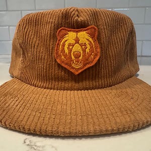May include: Brown corduroy baseball cap with a bear patch. The cap features a structured crown and a curved brim. The bear patch is in shades of orange and yellow, adding a contrasting detail to the hat.