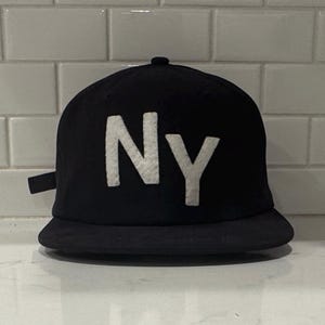 May include: Black baseball cap with a flat brim. The cap features the white letters "NY" on the front. The hat is made of a dark fabric and has a small, dark-colored brim.