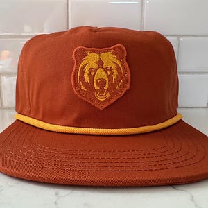 May include: A rust-coloured baseball cap with a yellow rope brim and a bear patch. The patch features a gold bear head design. The hat has a structured crown and a curved brim, suitable for outdoor activities.