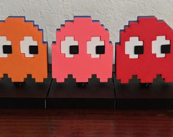 May include: A set of five pixelated Pac-Man characters in blue, orange, pink, red, and yellow. Each character is mounted on a black base. The yellow Pac-Man is open-mouthed, while the others are ghosts with white eyes and black pupils.
