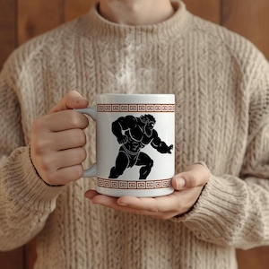 May include: A white ceramic mug featuring a black silhouette illustration of a muscular bearded figure in a dynamic pose. The design is framed by horizontal bands of brown Greek key patterns at the top and bottom of the mug, which contains hot liquid.