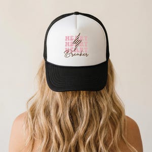 Puede incluir: Gorra de camionero en blanco y negro con un panel frontal blanco con el texto "HEART BREAKER" en rosa y un rayo gráfico. La gorra tiene una parte trasera de malla negra y una visera curva.