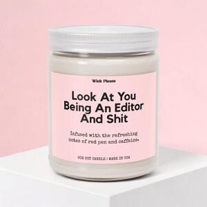 May include: A soy candle in a clear glass jar with a white lid. The pink label reads "Look At You Being An Editor And Shit." The candle is infused with red pen and caffeine notes. The jar is 9 oz.