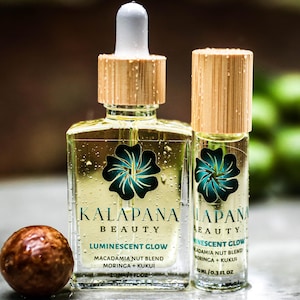 May include: Two glass bottles of Kalapana Beauty Luminescent Glow oil with wooden caps. The larger bottle is square, the smaller is cylindrical. Both have a teal flower design and the words "Kalapana Beauty" and "Luminescent Glow" printed on them. A round, brown nut is in the foreground.