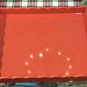 May include: A rectangular, coral-colored tray with scalloped edges and cut-out handles. The tray has a glossy finish and is likely made of wood or a similar material. It sits on a plaid surface.
