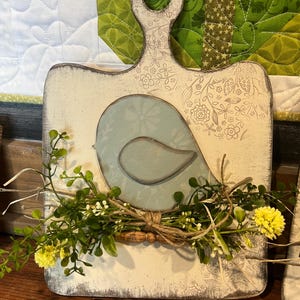 May include: A decorative wooden cutting board with a distressed white finish. It features a light blue bird design and is adorned with a twine-tied bundle of greenery and yellow flowers. The handle has a hole for hanging.