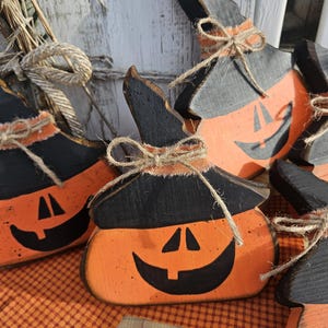 May include: A collection of orange and black wooden pumpkin decorations, each shaped like a pumpkin wearing a pointed witch hat. The pumpkins feature hand-painted black jack-o'-lantern faces and are accented with a tied jute twine bow.