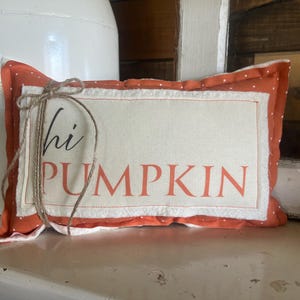 May include: A rectangular orange pillow with white polka dots featuring a cream-colored center panel with the text "hi PUMPKIN" in black and orange lettering. A thin brown twine bow is tied around the left side of the fabric pillow.