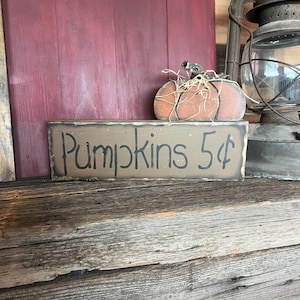 May include: A rectangular wooden sign with a distressed brown finish featuring the black hand-painted text Pumpkins 5¢. Resting on top is a small, flat, orange wooden pumpkin cutout adorned with a decorative bundle of dried raffia straw.