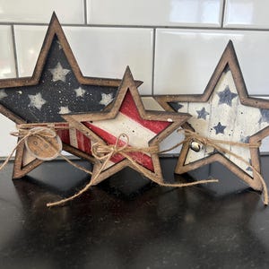 May include: Three wooden star decorations with a rustic, patriotic theme. Each star is painted with a different design: one with a blue background and white stars, one with red and white stripes, and one with a white background and blue stars. They are tied together with twine.