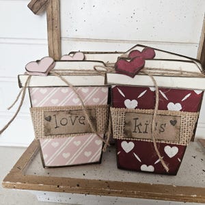 May include: Two decorative wooden coffee cup-shaped containers. One is pink with white hearts and the word "love". The other is burgundy with white hearts and the word "kiss". Both have burlap bands and heart-shaped lids.