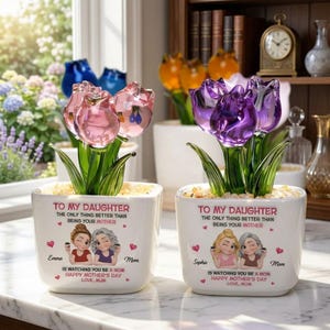 Op de afbeelding: Twee witte vierkante plantenbakken met kristallen tulpenbloemen. Eén heeft roze tulpen, de andere paars. Elke plantenbak heeft de tekst "TO MY DAUGHTER" en een Moederdagboodschap. De plantenbakken zijn versierd met cartoon illustraties.