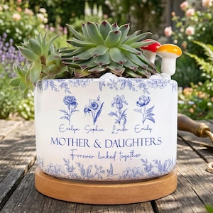 Puede incluir: Una maceta de cerámica blanca con diseños florales azules y el texto "MOTHER & DAUGHTERS Forever linked together." Contiene una planta suculenta y figuras de hongos. La maceta descansa sobre una base de madera, ideal para la decoración del hogar.
