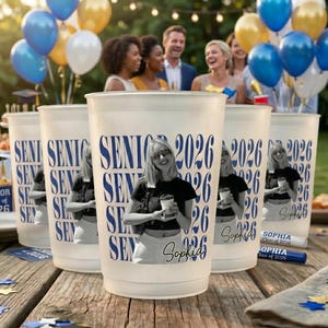 Pode incluir: Copos de pl&aacute;stico fosco com "SENIOR 2026" impresso em azul, com uma foto de uma pessoa. Os copos est&atilde;o em uma mesa de madeira, com bal&otilde;es ao fundo. Tema de festa de formatura.