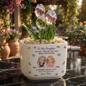 May include: A white ceramic planter with a Mother's Day message and cartoon illustrations. The planter holds glass tulip sculptures with green stems and clear, pink-tinted flower heads. The planter is on a black marble table.
