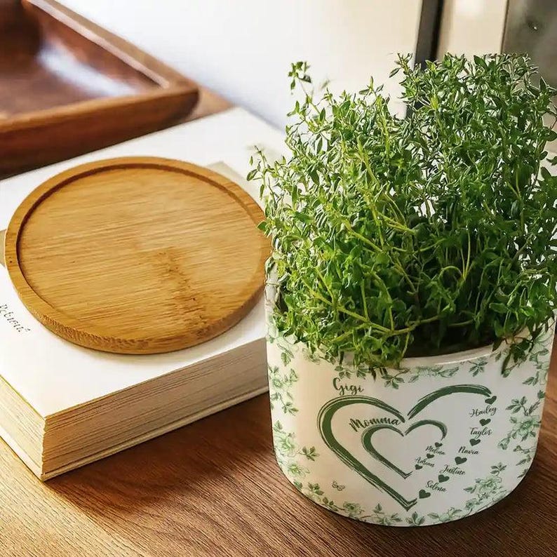 Peut inclure: Un plant de thym en pot dans un pot en c&eacute;ramique blanc avec un motif de c&oelig;ur vert et les mots "Gigi Momma" et des noms. Un dessous de verre rond en bambou et une pile de livres sont &eacute;galement visibles.