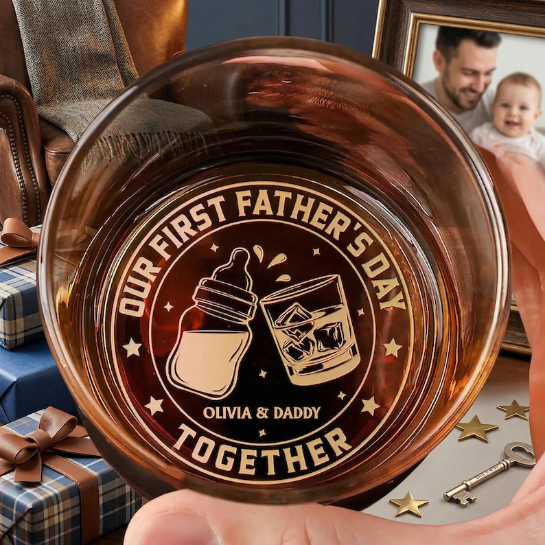 K&ouml;nnte beinhalten: Ein braun get&ouml;ntes Glas mit den Worten "OUR FIRST FATHER'S DAY TOGETHER" und einer Babyflasche und einem Whiskeyglas, die darauf ge&auml;tzt sind. Die Namen "OLIVIA & DADDY" sind ebenfalls auf dem Glas eingraviert. Das Glas wird von einer Person gehalten.