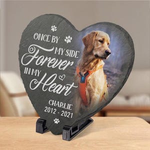 May include: Heart-shaped slate memorial stone with a photo of a golden retriever. The stone features the text "Once by my side, forever in my heart" and the name "Charlie 2012-2021". The stone is supported by a black stand.
