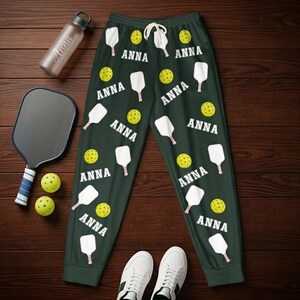 Könnte beinhalten: Dunkelgrüne Jogginghose mit weißem Kordelzug, mit einem Muster aus Pickleball-Schlägern, gelben Bällen und dem Namen "ANNA" in Weiß. Das Bild zeigt auch einen Pickleball-Schläger, Bälle, eine Wasserflasche und weiße Turnschuhe.