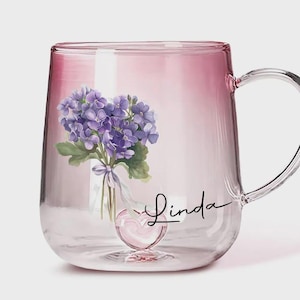 Könnte beinhalten: Transparente Glas-Tasse mit rosa Farbverlauf. Die Tasse hat ein Blumendesign aus lila Blumen und den Namen "Linda" in Schreibschrift. Ein herzförmiges Detail befindet sich am Boden der Tasse.