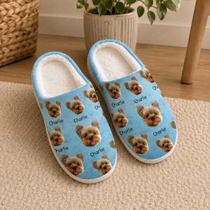 May include: A pair of light blue house slippers featuring a repeating pattern of a Yorkshire Terrier's face and the name "Charlie". The interior is white and fluffy.