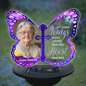 May include: A solar-powered memorial garden stake in the shape of a butterfly. The butterfly has a photo of a woman, the years 1935-2024, and the text "Your Wings were ready but my Heart was not." The stake is purple and white.
