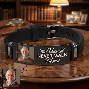 Può includere: Bracciale in pelle nera con fibbia e dettagli argentati. Il bracciale presenta una placca rettangolare nera con la foto di un uomo e la scritta bianca "You Never Walk Alone".