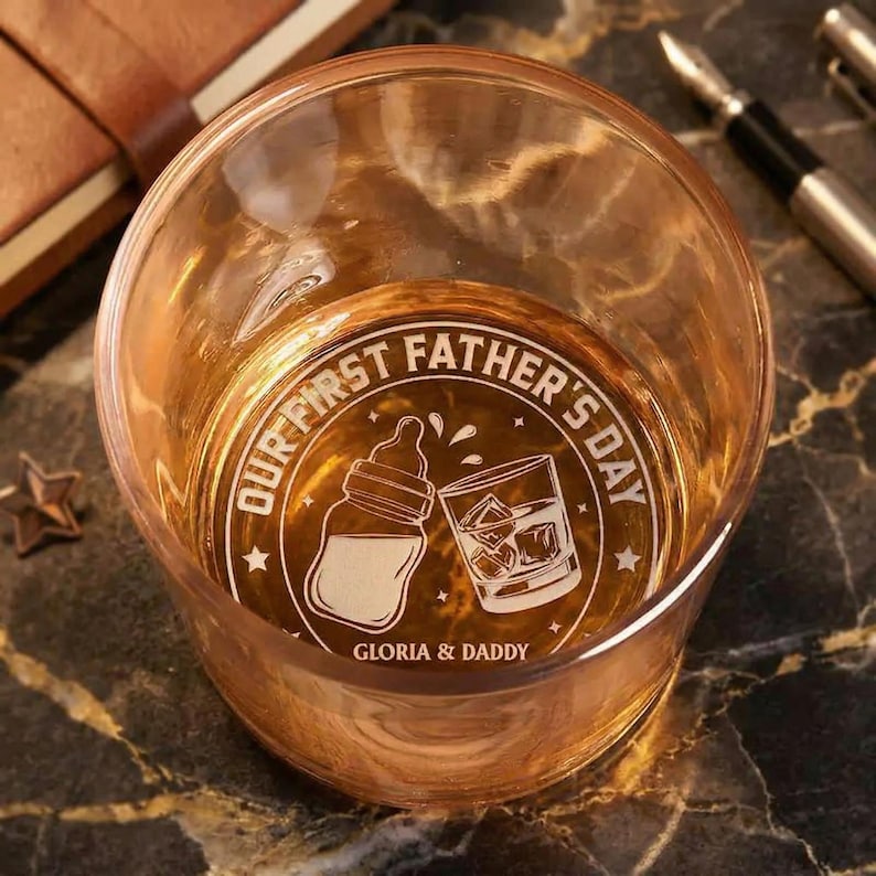 K&ouml;nnte beinhalten: Ein klares, rundes Glas mit Goldt&ouml;nung, mit einer wei&szlig;en Grafik mit der Aufschrift "OUR FIRST FATHER'S DAY" mit einer Babyflasche und einem Glas Whisky. Der Text "GLORIA & DADDY" befindet sich darunter. Stifte und ein Notizbuch im Hintergrund.
