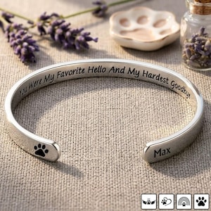 Könnte beinhalten: Ein silbernes Armreif-Armband mit der Inschrift "You Were My Favorite Hello And My Hardest Goodbye." Das Armband hat einen Pfotenabdruck und den Namen "Max." Es liegt auf einer strukturierten Oberfläche, mit Lavendel und einer pfotenförmigen Schale.