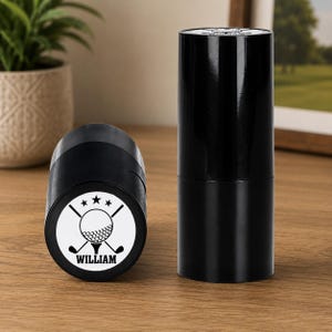 May include: Two black cylindrical containers. One is lying on its side, displaying a white circular emblem with a golf ball, crossed golf clubs, three stars, and the name "WILLIAM". The other container stands upright.