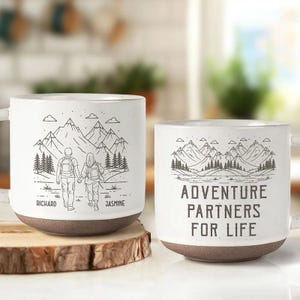 May include: Two white ceramic mugs with brown bases. One mug features a mountain scene with a couple and the names "Richard" and "Jasmine." The other mug has the text "ADVENTURE PARTNERS FOR LIFE" with a mountain range.