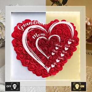 Pu&ograve; includere: Una composizione floreale a forma di cuore in una cornice bianca, piena di rose rosse. Il testo bianco recita "Nonna" e "Mamma". Ulteriori nomi sono elencati di seguito. La cornice ha una funzione di luce LED, con opzioni "OFF" e "ON".