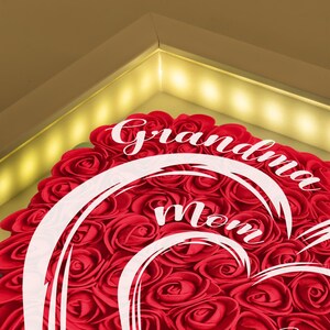 Pu&ograve; includere: Una composizione a forma di cuore di rose rosse con le parole "Nonna Mamma" in corsivo bianco. Le rose sono fittamente disposte, creando una superficie strutturata. La composizione &egrave; incorniciata con un'illuminazione calda.