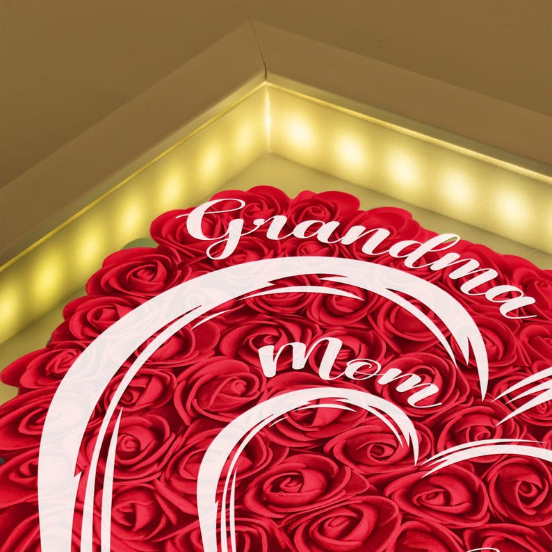 Pu&ograve; includere: Una composizione a forma di cuore di rose rosse con le parole "Nonna Mamma" in corsivo bianco. Le rose sono fittamente disposte, creando una superficie strutturata. La composizione &egrave; incorniciata con un'illuminazione calda.
