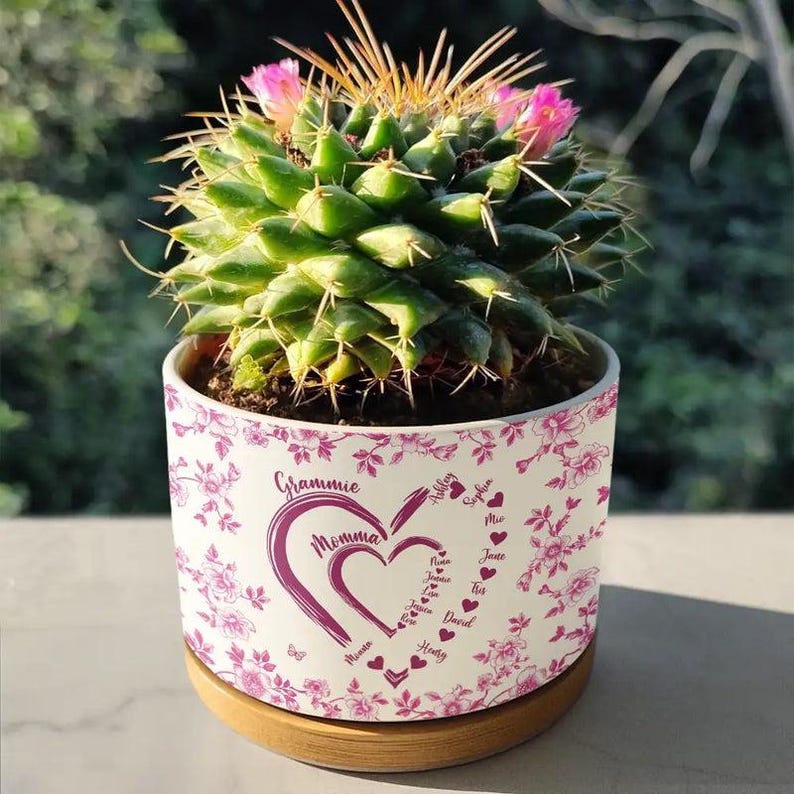 Peut inclure: Un cactus vert avec des fleurs roses dans un pot en c&eacute;ramique blanc orn&eacute; d'un motif floral et des mots "Grammie Momma" dans un c&oelig;ur, entour&eacute; de noms. Le pot repose sur une soucoupe en bois. Le diam&egrave;tre du pot est d'environ 15 cm.