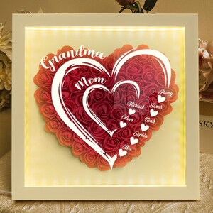 Pu&ograve; includere: Una composizione floreale a forma di cuore incorniciata con le parole "Grandma" e "Mom" in scrittura bianca. Il cuore &egrave; pieno di rose rosse e include nomi. La cornice ha una luce calda e brillante.