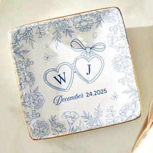 Può includere: Piatto quadrato in ceramica bianca con motivo floreale blu e bordo dorato. Presenta due medaglioni a forma di cuore con le iniziali "W" e "J", e la data "24.12.2025". Piatto decorativo per occasioni speciali.