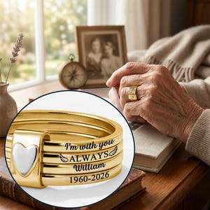 May include: Gold ring with a white heart-shaped detail. The ring is inscribed with the words "I'm with you ALWAYS William 1960-2026". The ring is on a person's finger. The ring is a memorial or remembrance jewelry.