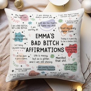 May include: A white square cushion featuring the words "EMMA'S BAD BITCH AFFIRMATIONS" in black. The cushion is decorated with various affirmations in different fonts and colours, such as "I am a boss, ruling my world" and "I'm an Unstoppable Fucking Force of Nature."