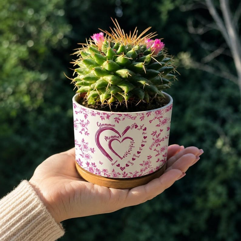 Peut inclure: Un petit cactus avec des fleurs roses dans un pot en c&eacute;ramique blanc. Le pot a un motif floral avec les mots "Grammie" et "Momma" en forme de c&oelig;ur. Le pot repose sur une soucoupe en bois.