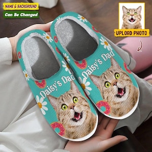 May include: Teal slippers with a smiling cat face, daisies, and donuts. The text "Daisy's Dad" is printed on the side. The slippers have a soft, grey interior.
